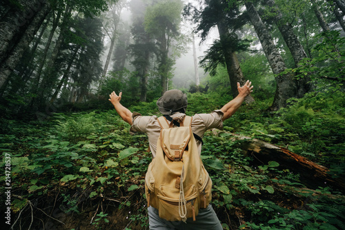 a male tourist in shorts, a hat and with a beige backpack in the fog forest of the hand to the sides, a gesture of admiration.