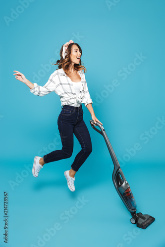 Full length photo of funny housewife 20s listening to music via wireless headphones and jumping with vacuum cleaner, isolated over blue background