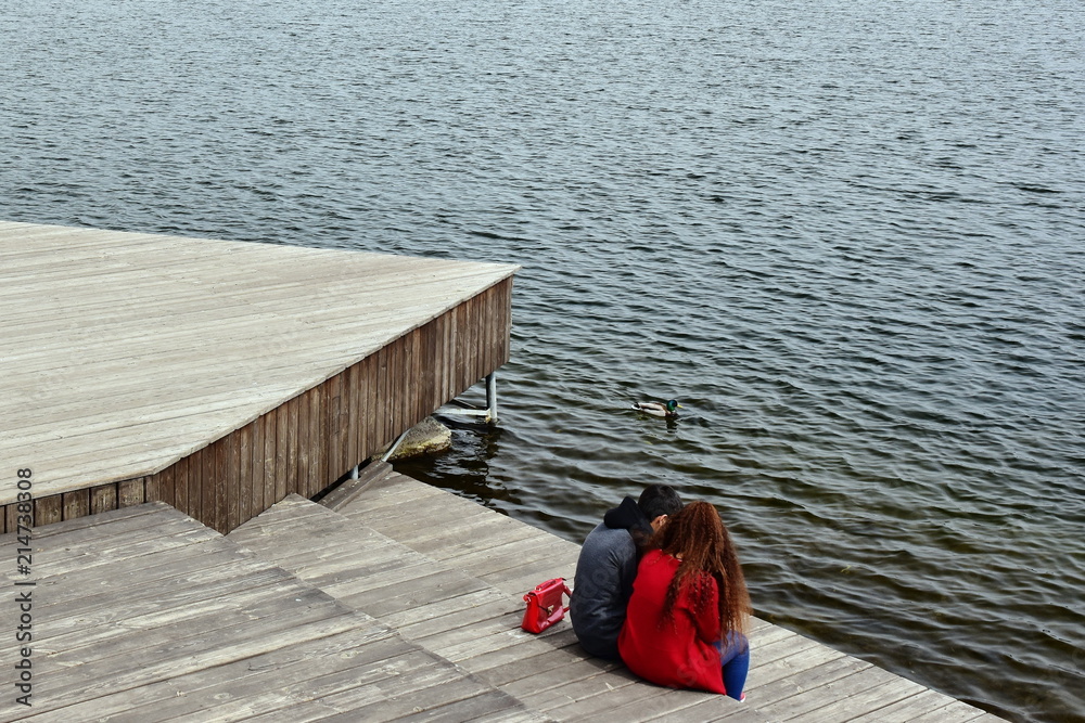 Love warms in cold weather.loving couple sitting on waterfront on wooden boards.They look at the river.girl in red,red curly hair.guy in black jacket Love warms in cold weather.loving couple sitting on waterfront on wooden boards.They look at the river.girl in red,red curly hair.guy in black jacket