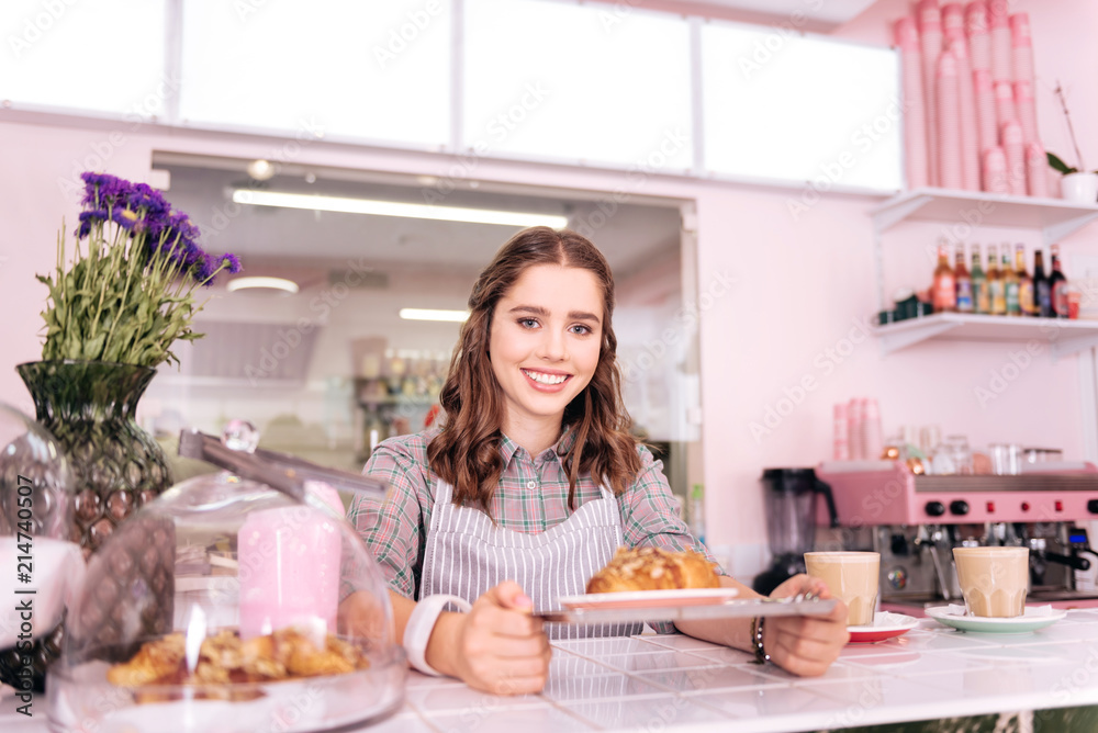Young barista. Appealing young barista smiling broadly while preparing two latte and one croissant