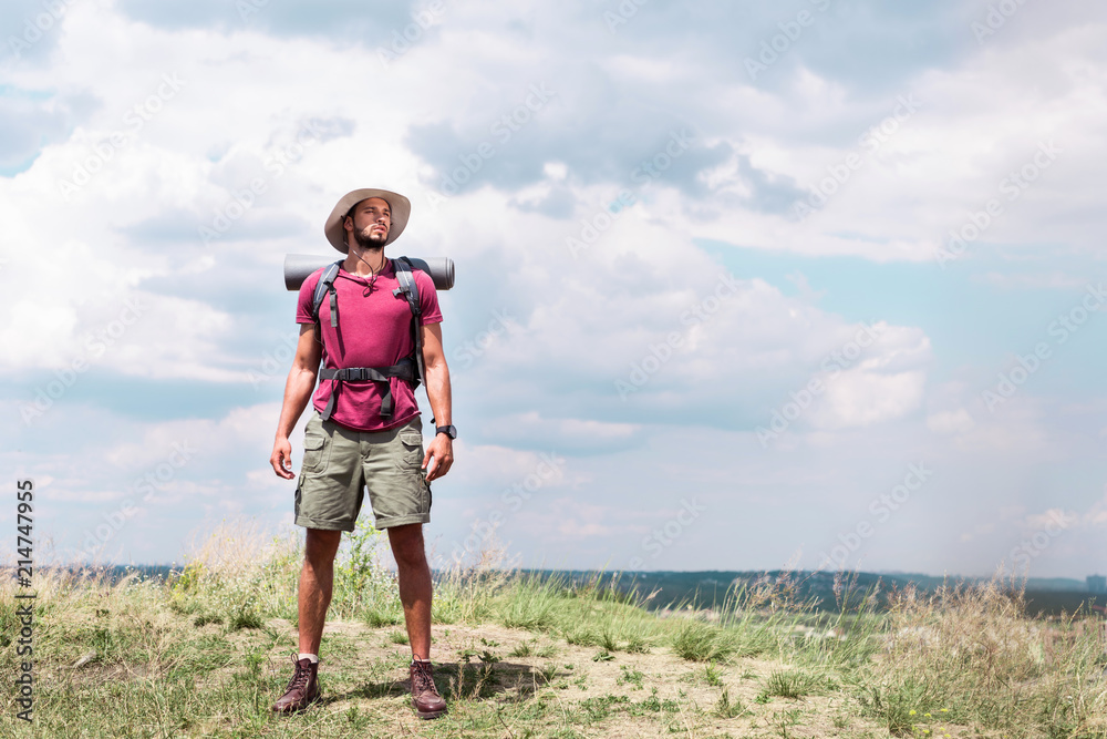 handsome hiker with backpack standing on summer meadow with cloudy sky