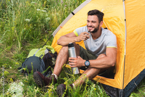 smiling traveler sitting in yellow tent with backpack and drinking coffee from thermos
