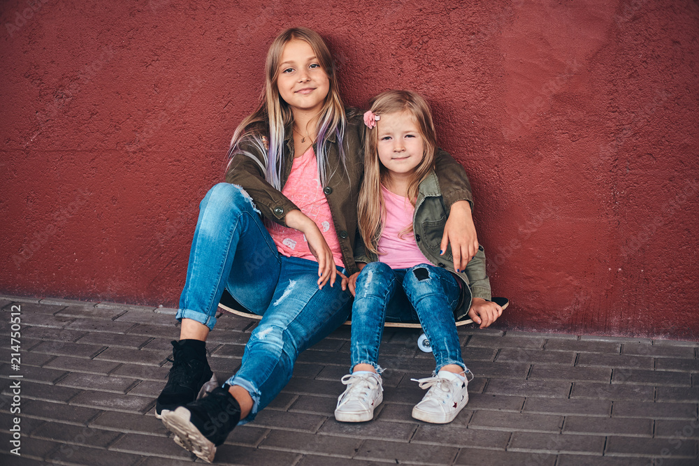 Portrait of two little sisters dressed in trendy clothes leaning on a wall while sitting on a skateboard at a bridge footway. Portrait of two little sisters dressed in trendy clothes leaning on a wall while sitting on a skateboard at a bridge footway.
