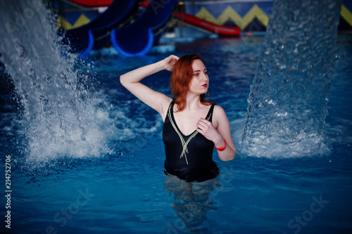 Sexy red haired girl in black swimsuit having rest in swimming pool of aqua park.