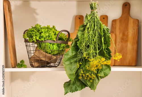 Hang-up bunch of herbs near basket with fresh lettuce on shelf