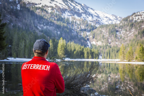 hiker with red sweater with the word österreich on it is standing in front of lake in austria