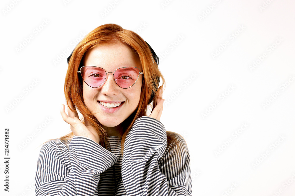 Young attractive natural redhead woman smiling dancing, enjoying favorite song with large headphones. White background, copy space. Attractive red hair female in casual outfit listening beloved tune.