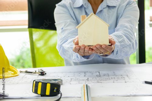 Engineering woman holding  wooden house model with architecture blueprint and equipment tools background. Selective focus.