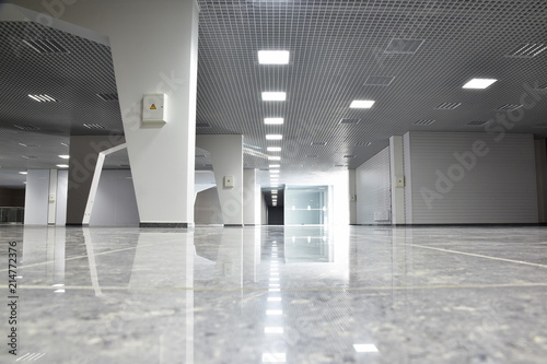 Unfinished building interior, white hall. The premise of the shopping center after repair. On the floor, granite tiles reflect light from the fixtures. Drywall arches. Grilyato ceiling