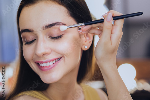 Everyday makeup. Pleasant smiling makeup artist feeling joyful while finishing her everyday makeup