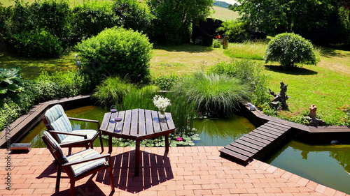 attraktiver naturbelassener Teich an rote Terrasse angrenzend mit Brücke und Tisch und Stühlen