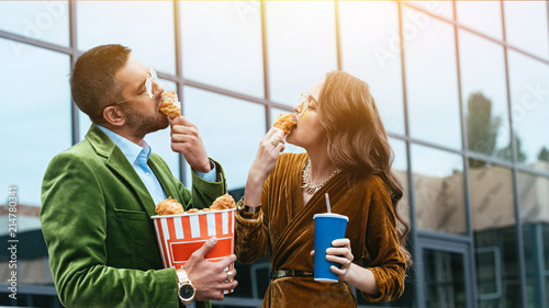 side view of fashionable couple in velvet clothing eating fried chicken legs on street