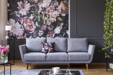 Grey lounge with patterned cushion in real photo of dark living room interior with floral wallpaper, molding on wall and gold lamp