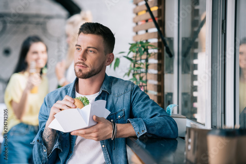 handsome man holding burger and looking away near food truck
