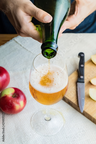 Close-up image of man pouring premium cidre in glass. Top view of male hands pouring vintage apple wine into beautiful glass in rustic table background with ripe apples photo