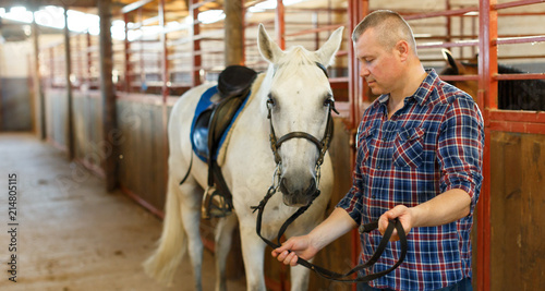 Positive man farmer with white horse standing at stabling indoor