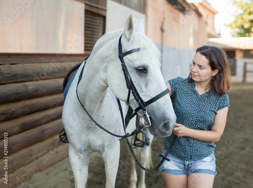 Mature smiling woman farmer standing with white horse at stable