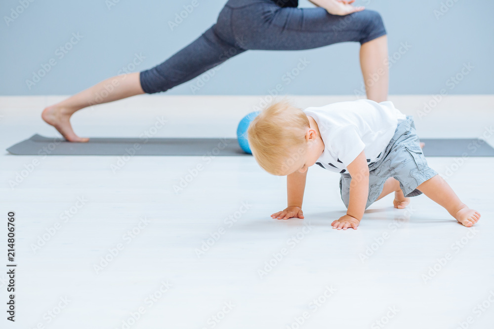 Naklejka premium Beautiful mother and little toddler son practicing yoga together at home, making stretching exercise, copy space. Kids crawling on foreground.