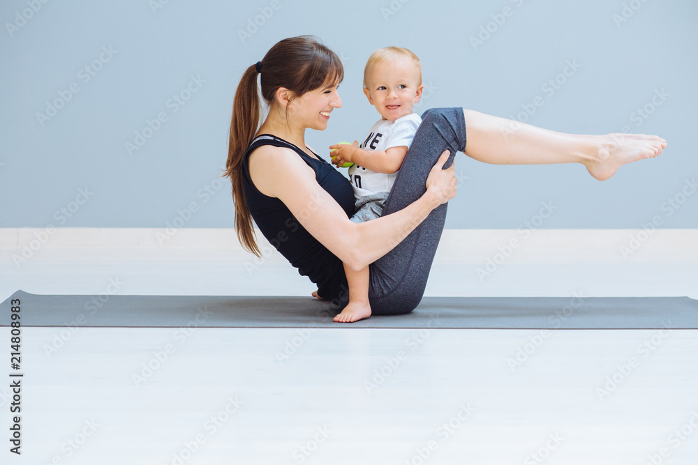 A sports mother is engaged in fitness and yoga or pilates exercie with a toddler son at home. Empty gray background. Motherhood, simple healthy lifestyle concept. A sports mother is engaged in fitness and yoga or pilates exercie with a toddler son at home. Empty gray background. Motherhood, simple healthy lifestyle concept.