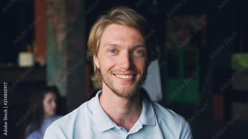 Close-up slow motion portrait of handsome bearded man company employee looking at camera and smiling while his coworkers are working in background.