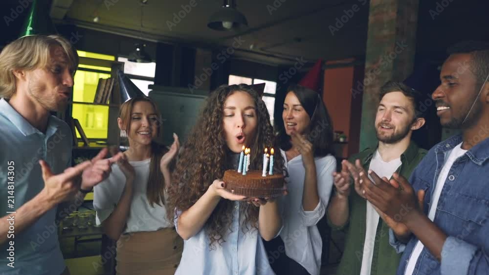 Slow motion of beautiful girl in party hat holding cake and blowing candles while coworkers are standing around, clapping hands and congratulating her in office.