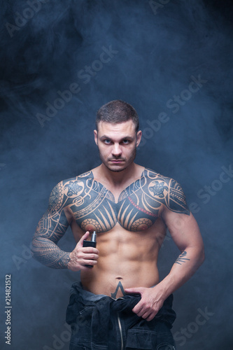 Vaper. The man with a muscular naked torso with tattoos smoke an electronic cigarette on the dark background