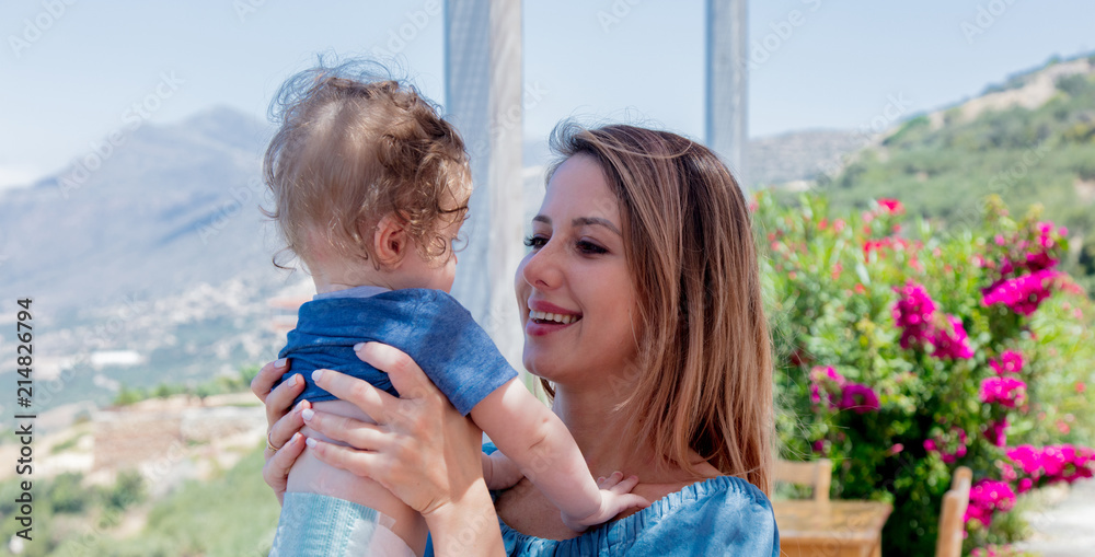 Fototapeta premium Young caucasian mother with a child have a vacation on Crete, Greece. Mother hold a boy on hands and landscape of a summertime season flowers and olive trees