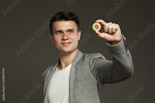 portrait of a young guy in a jacket with a bitcoin coin in his hand on a gray background