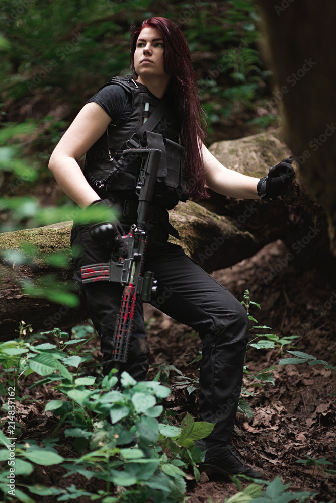 Young woman with gun in hands in dark uniform among trees.Selective focus