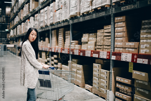 Half length portrait of beautiful young asian woman with empty cart looking for something at self service warehouse. Shopping concept
