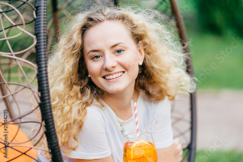 Horizontal shot of pretty smiling European female has wavy bushy light hair  holds fresh cocktail  sits on hanging chair  breathes fresh air. Positive emotions  recreation and people concept