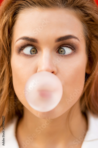 Funny portrait of beautiful young cute woman in white vogue suit look to her nose while blowing gum bubble against red wall. Urban style girl, quick lifestyle and work concept. Hipster city life.