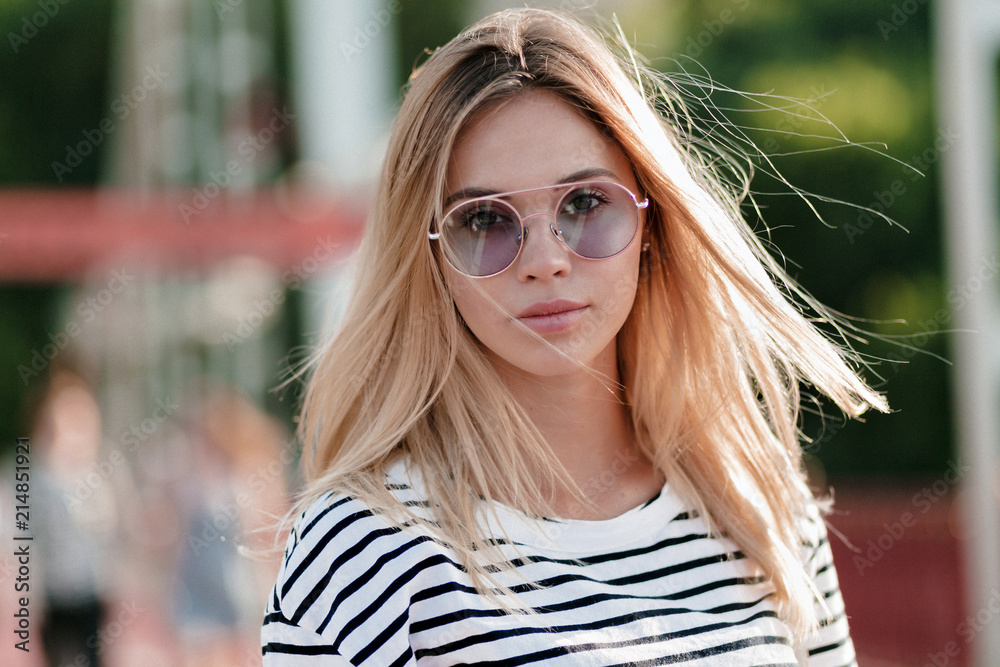 Close-up outdoor portrait of lovely adorable young woman with blond hair wears stylish bright glasses and stripped t-shirt walking on the bridge in sunlight Close-up outdoor portrait of lovely adorable young woman with blond hair wears stylish bright glasses and stripped t-shirt walking on the bridge in sunlight