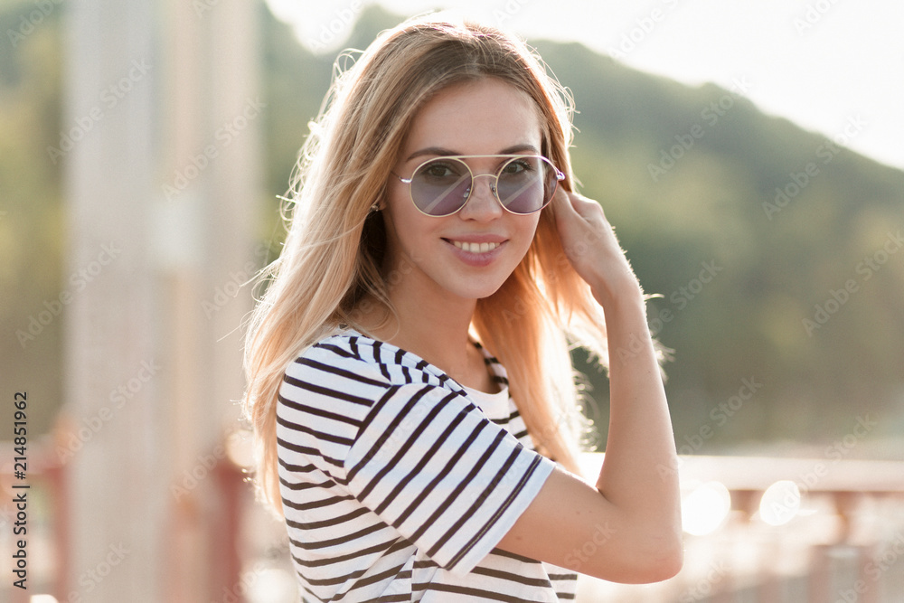Outdoor shot of glad friendly woman wearing striped attire and round violett glasses. Smiling blonde woman posing with lovely smile in sunny warm day