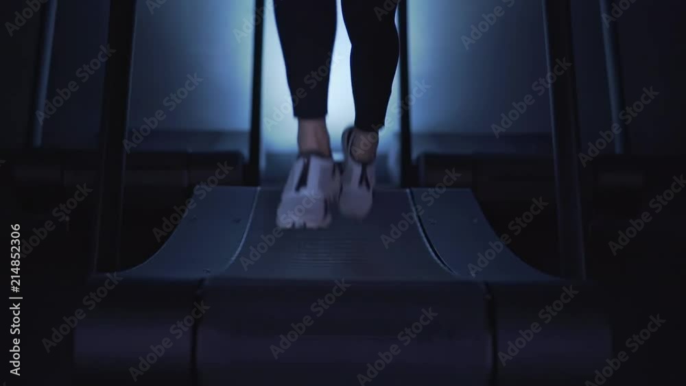 Woman s legs in sneakers walking then running on a treadmill in a dark gym. Locked down slow motion medium shot