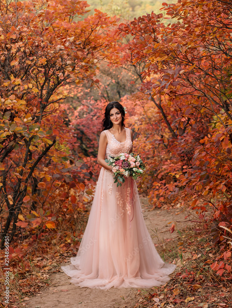 Girl brunette with long hair, in a luxurious pink dress with a long train. The bride with a bouquet poses against a background of a fabulous autumn landscape, in red and orange shades. Art film photo