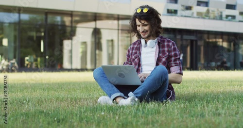 Caucasian young man in the motley shirt typing on his laptop computer and sitting on the grass near his college. Outside. photo