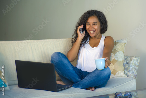 lifestyle isolated portrait of young happy and gorgeous black african American woman talking on mobile phone while working on laptop computer at home couch
