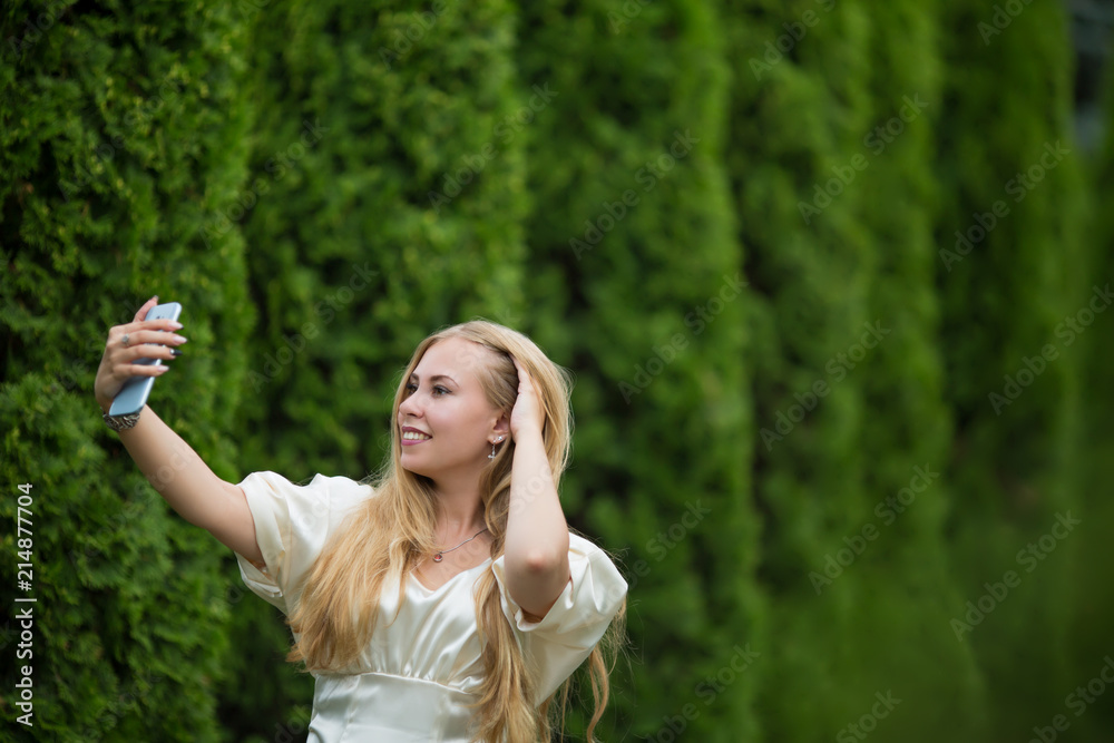 Young pretty blonde girl in a dress makes a selfie between thuja on the street in summer.