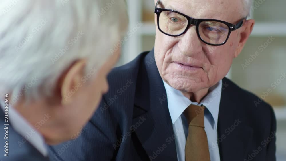 Tilt up of serious senior businessman in suit and glasses discussing work with his male elderly colleague during meeting in office Tilt up of serious senior businessman in suit and glasses discussing work with his male elderly colleague during meeting in office