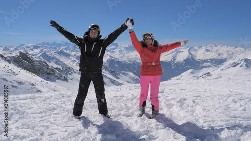 Happy skiers couple stands on the mountain peak and rises their hands