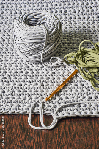 skein of grey knitted yarn and a crochet hook against the background of a knitted carpet