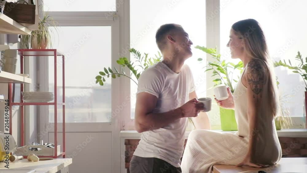 The pretty tattooed smiling lovers in white are having a dialogue while drinking coffee and tea The pretty tattooed smiling lovers in white are having a dialogue while drinking coffee and tea