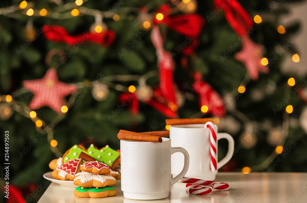 Cups of delicious hot cocoa with cinnamon sticks and cookies on table against blurred Christmas tree Cups of delicious hot cocoa with cinnamon sticks and cookies on table against blurred Christmas tree