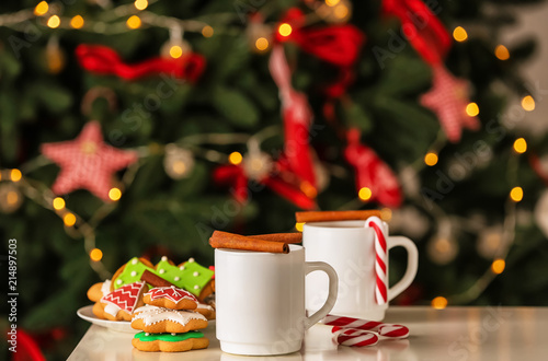 Cups of delicious hot cocoa with cinnamon sticks and cookies on table against blurred Christmas tree