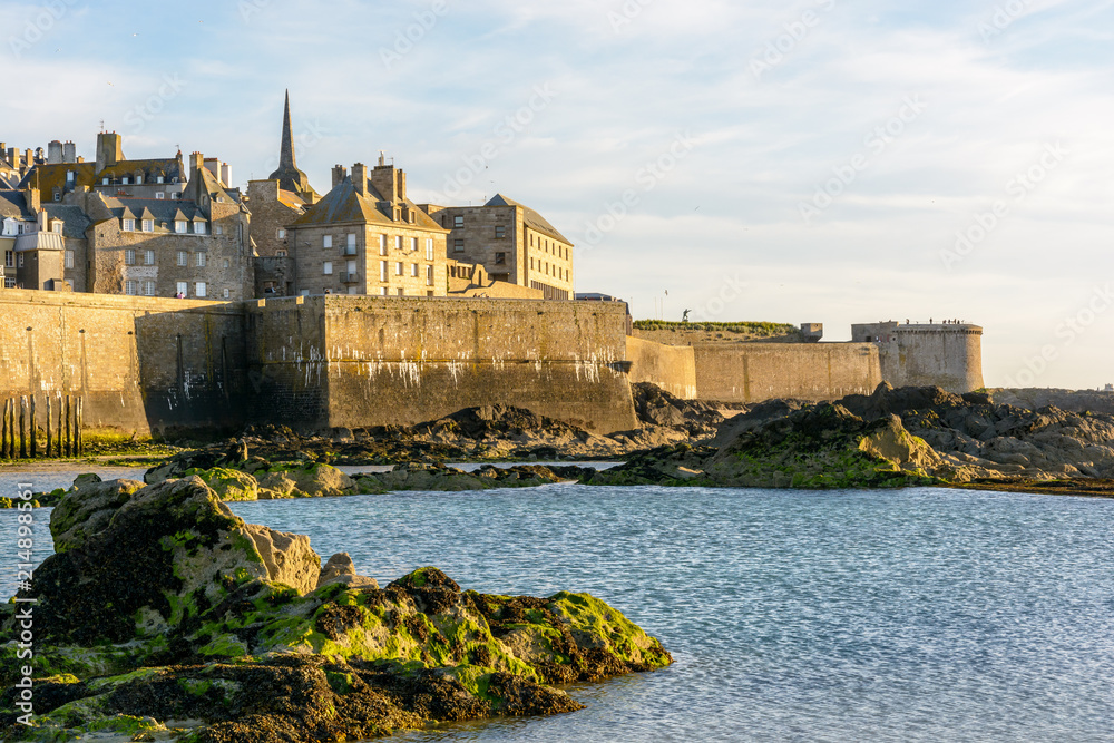 The walled city of Saint-Malo, France, at sunset with a spire sticking out from the buildings behind the wall, the Fort a la Reine, the Bidouane tower and a rock in the sea in the foreground.