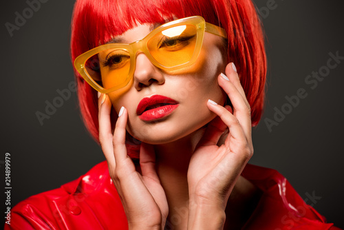 close-up portrait of seductive young woman in red latex jacket and vintage yellow sunglasses isolated on grey