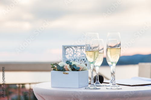 Wineglasses of ice champagne on table for wedding reception after wedding ceremony by the sea and sunset. Box with flowers with rings.