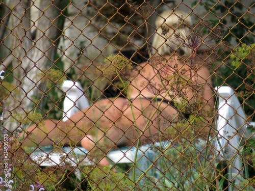 Voyeurism in summer, peeping on neighbor girls. View through the wire mesh fence on a sunbathing blonde girl in a bikini, selective focus