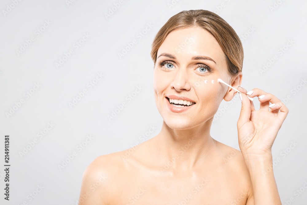 Portrait of young beautiful woman smiling while taking some facial cream using a cotton swab isolated on white background with copy space. Portrait of young beautiful woman smiling while taking some facial cream using a cotton swab isolated on white background with copy space.
